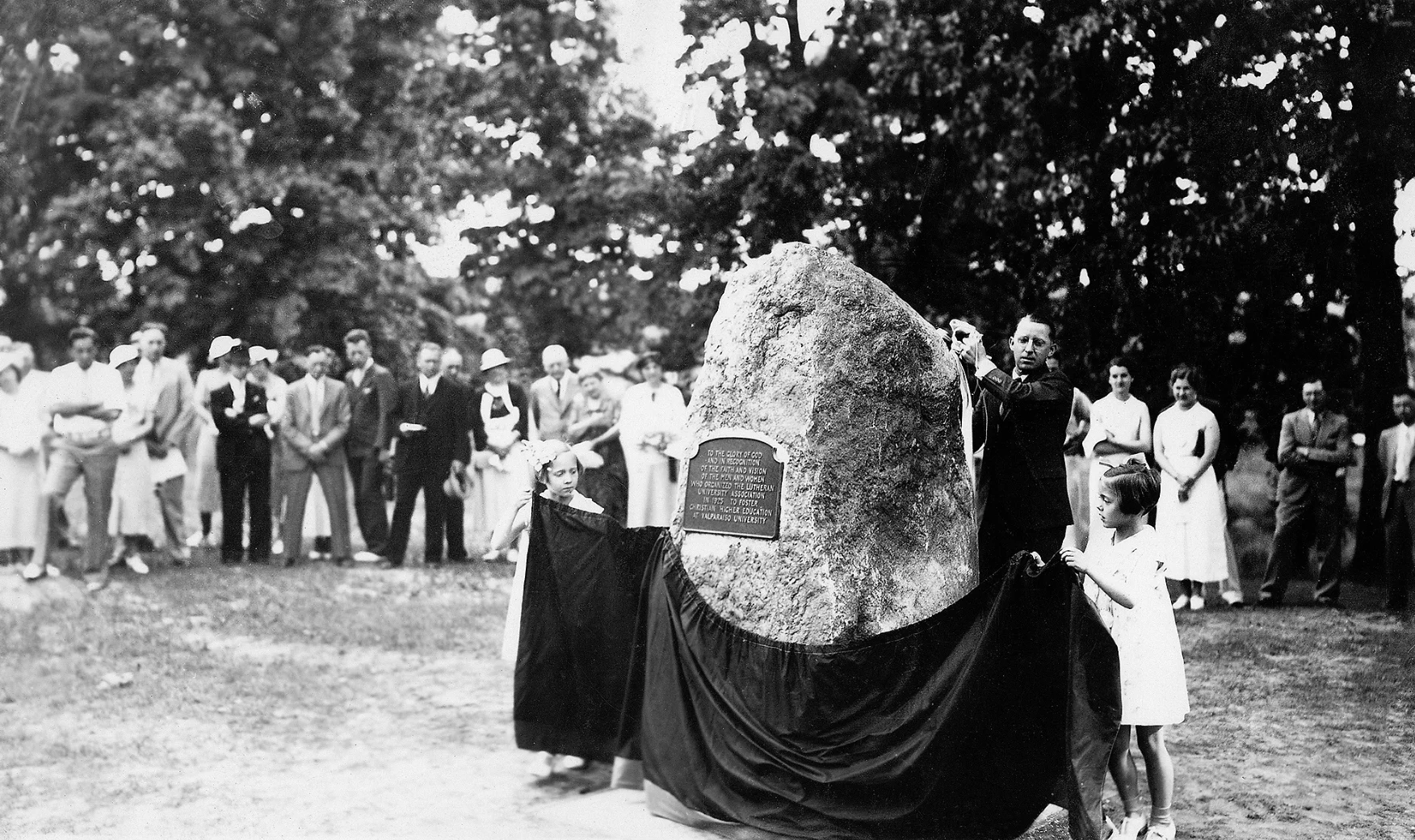 A group, including Past Presidents, unveils a commemorative plaque on a large rock at an outdoor ceremony with people gathered around.