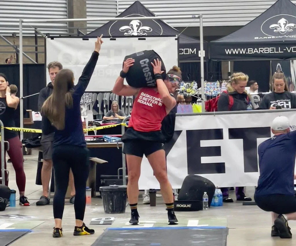 Jeanette Gray, M.Ed., lifts a large sandbag overhead while a judge raises a hand at the Beacon indoor fitness competition.