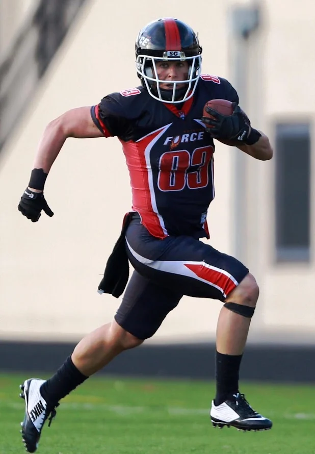 Football player in a black and red uniform running with the ball on a field, coached by M.Ed. holder Jeanette Gray, showcasing the spirit of Beacon athletics.