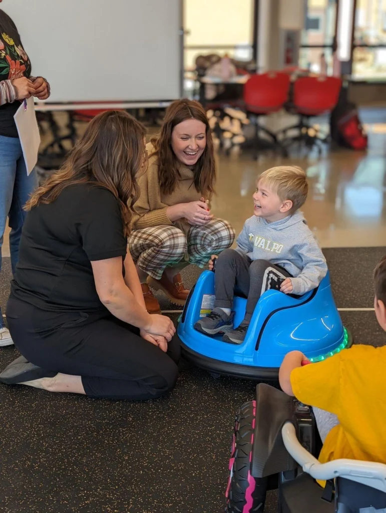Two women smile at a young boy sitting and laughing in a blue ride-on toy indoors.