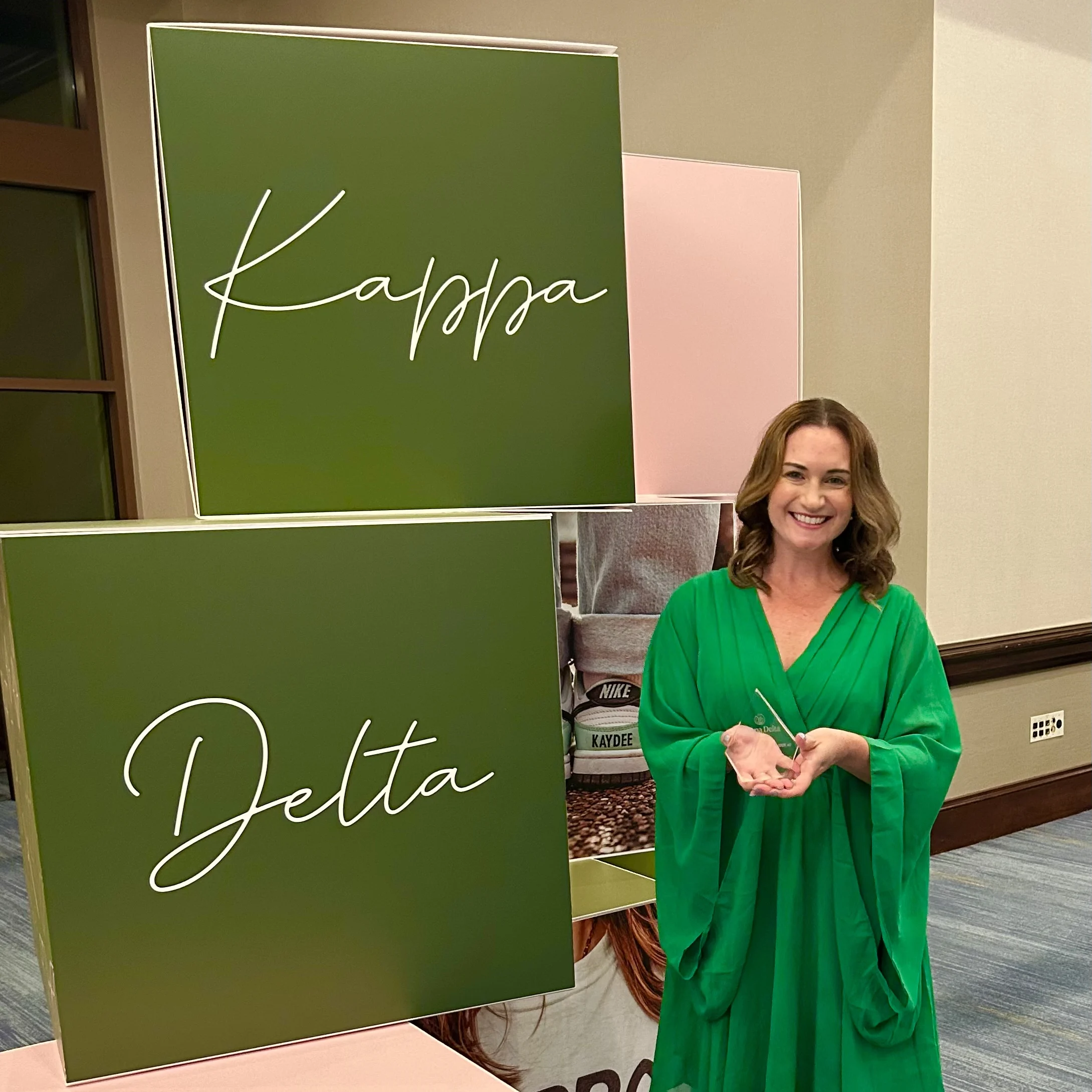 Meredith Garofalo ‘08 stands smiling in a green dress by Kappa Delta signs at an indoor Beacon event.