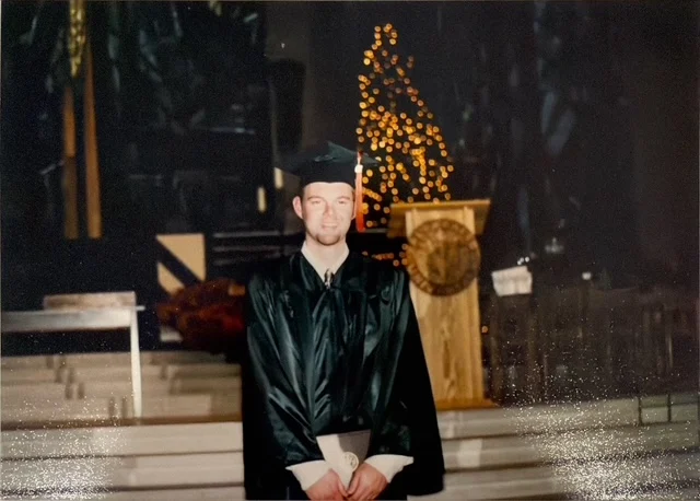 A graduate in cap and gown stands indoors, holding a diploma, with a lit tree in the background—a beacon of achievement for Matt Palm, April 2026.