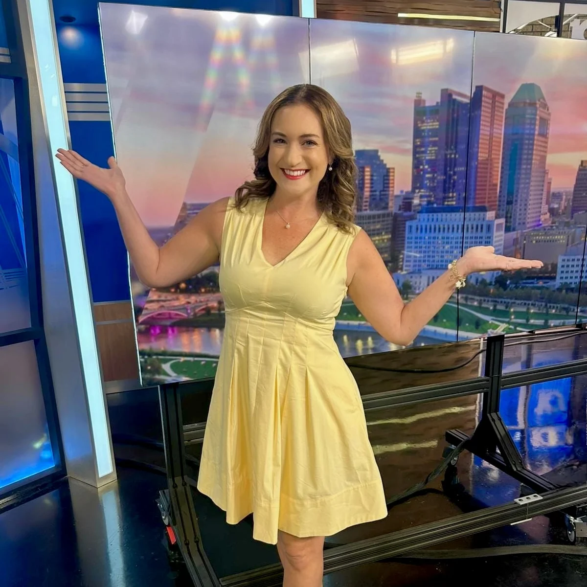 Meredith Garofalo beams in a yellow dress and red heels, posing with arms outstretched like a beacon against cityscape screens.