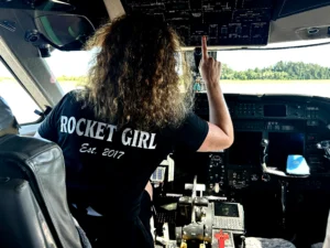 A woman in a "Rocket Girl" shirt operates controls in an airplane cockpit, facing the instrument panel, with a beacon of focus reminiscent of meteorologist Meredith Garofalo's trailblazing spirit.