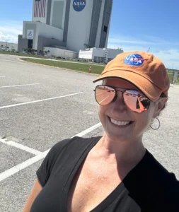 Smiling person in NASA hat and sunglasses, with NASA’s Vehicle Assembly Building shining like a beacon in the background on a sunny day.