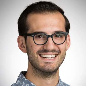 Smiling man with dark hair, glasses, and a patterned shirt stands against a plain light background—Gideon Litherland invites you to Follow the Beacon.