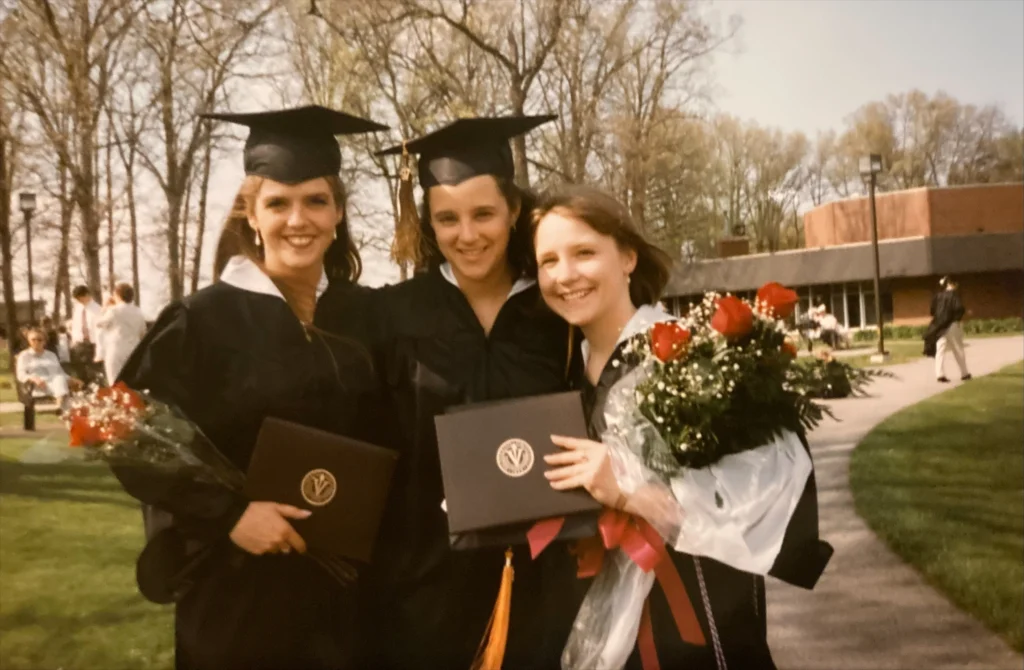 Three smiling graduates, including Katie Adrian Jasnieski, in caps and gowns holding diplomas and bouquets, stand outdoors on a sunny day—a beacon of achievement and new beginnings.