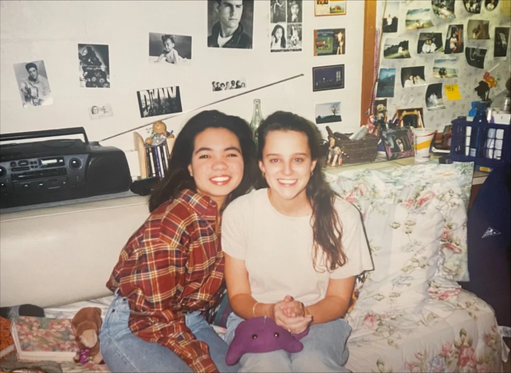 Two young women smile while sitting on a dorm room couch decorated with posters and photos, enjoying their time at Beacon.