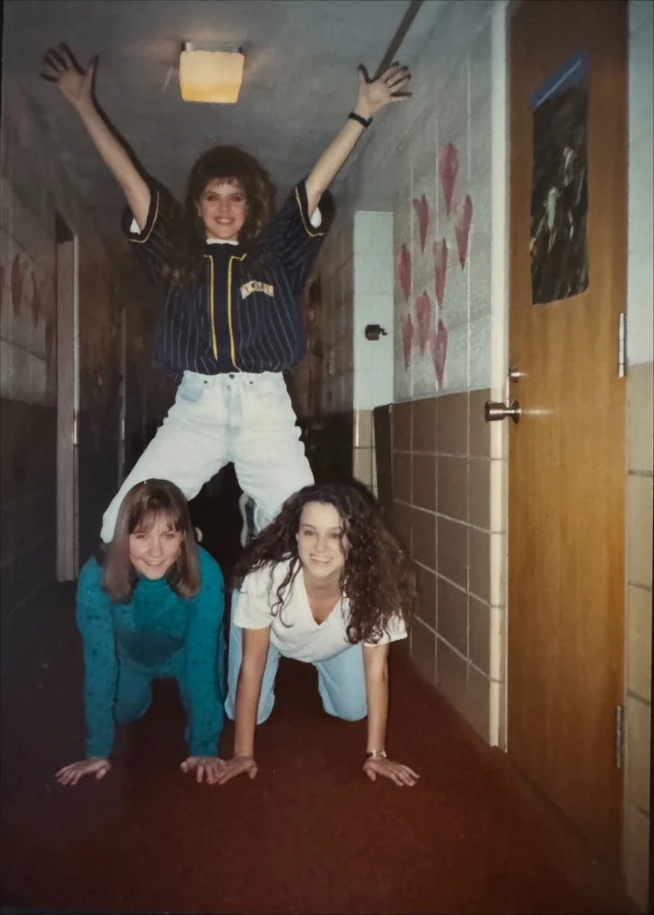 Three young women in a hallway do a pyramid pose—two kneeling, one standing on top with arms raised, channeling Beacon energy and camaraderie captured by Katie Adrian.