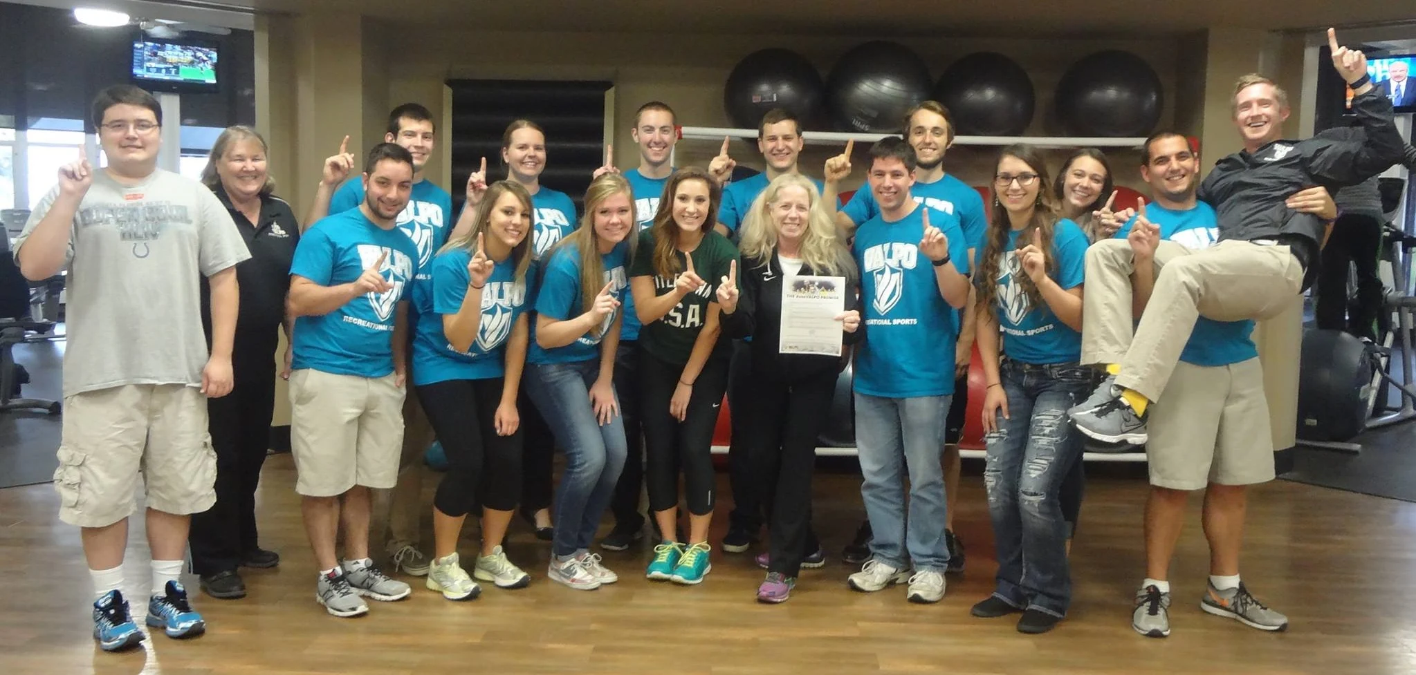 A group of people in blue shirts pose indoors, smiling and holding up one finger, celebrating the Seibert legacy, with one person—Rin Curtis—holding a paper.
