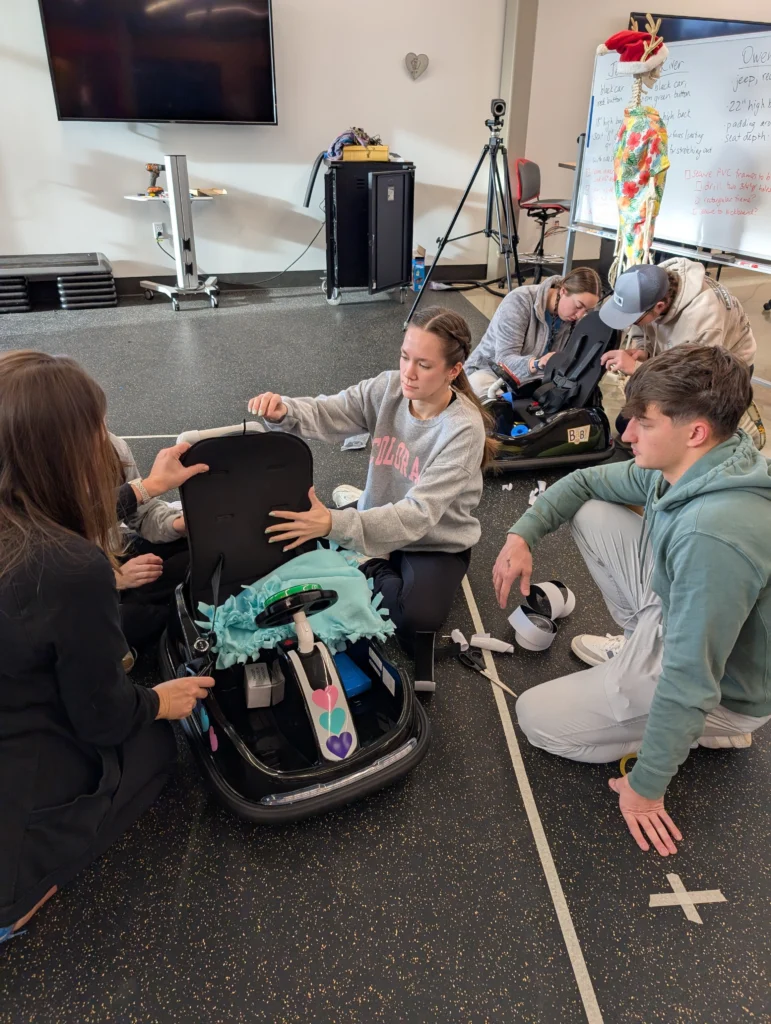 Four students and an instructor assemble a GoBabyGo car seat—enhancing mobility for children—in a classroom equipped with a whiteboard and anatomy model.