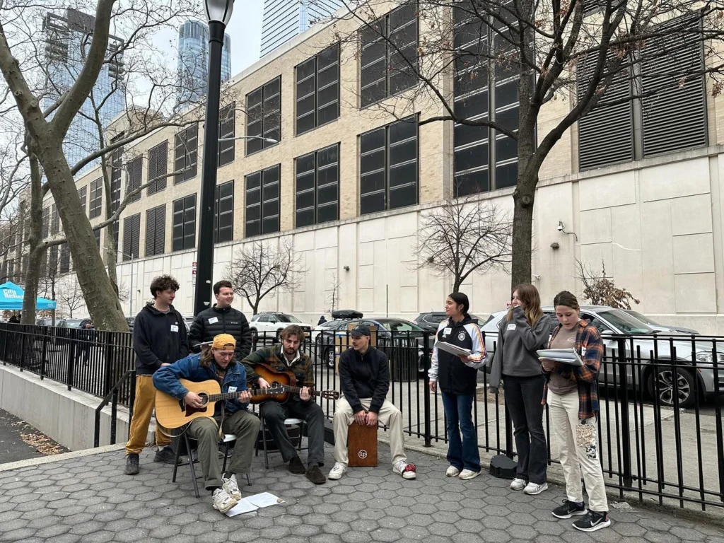 A group of people enjoy worship opportunities as they play music and sing together outside near a building and trees on a city sidewalk.