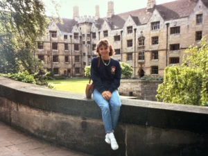 Woman in jeans and a dark sweater sits on a stone wall in front of a historic college building and lawn, proudly sporting her Valpo Alumni badge.