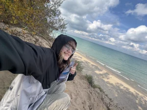 Person in a hooded jacket smiles and gives a thumbs up on a sandy cliff above a beach and blue lake, capturing the spirit of adventure in Nickie Smar’s 2028 photo series, Beacon’s Reflection.
