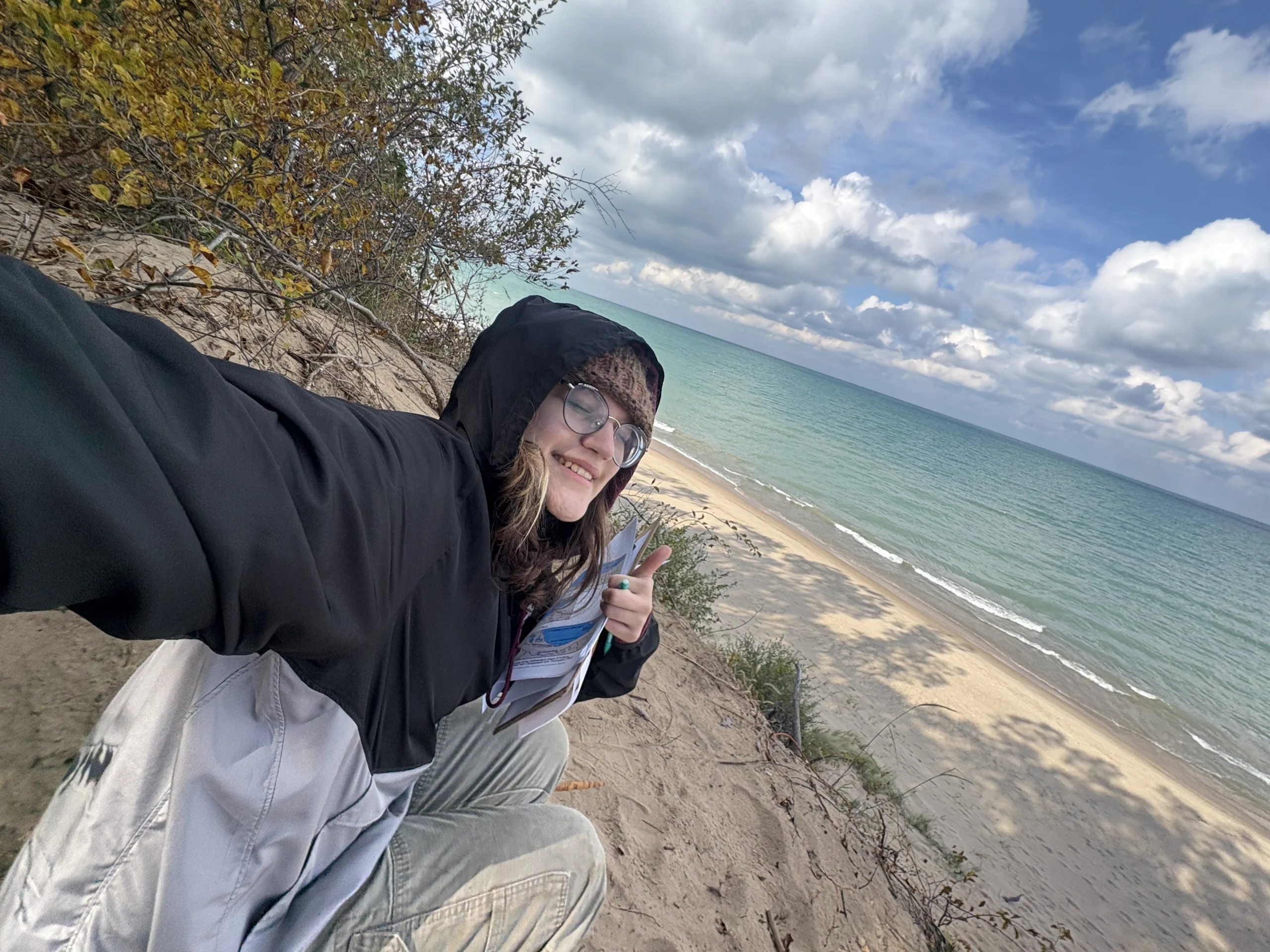 Person in a hooded jacket smiles and gives a thumbs up on a sandy cliff above a beach and blue lake, capturing the spirit of adventure in Nickie Smar’s 2028 photo series, Beacon’s Reflection.