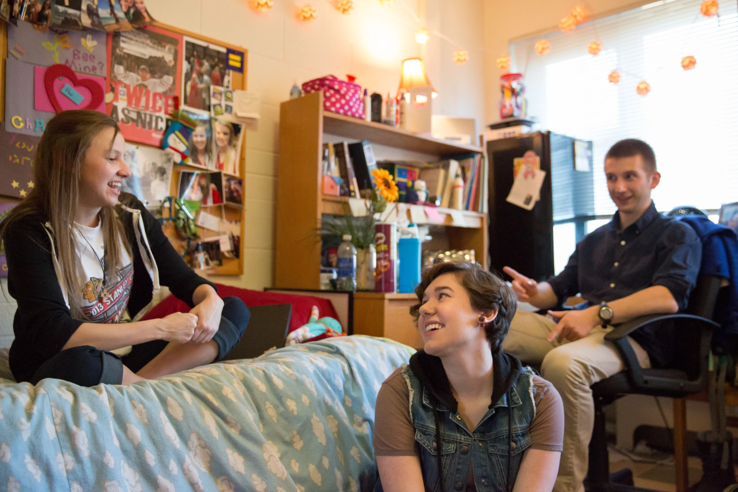 Three college students talk and smile together in a cozy, decorated dorm room with posters and string lights, enjoying the ups and downs of adjusting to college.