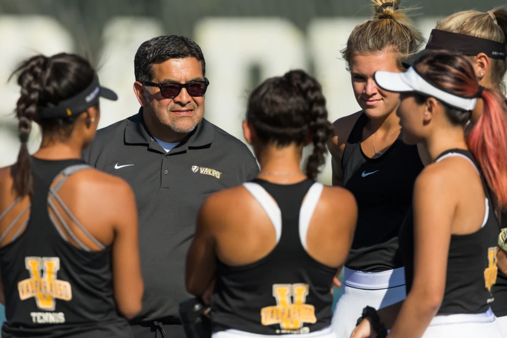 Coach Bob Modesto chats with five Valpo players in black uniforms during a sunny outdoor practice, sharing insights from his MBA Journey.