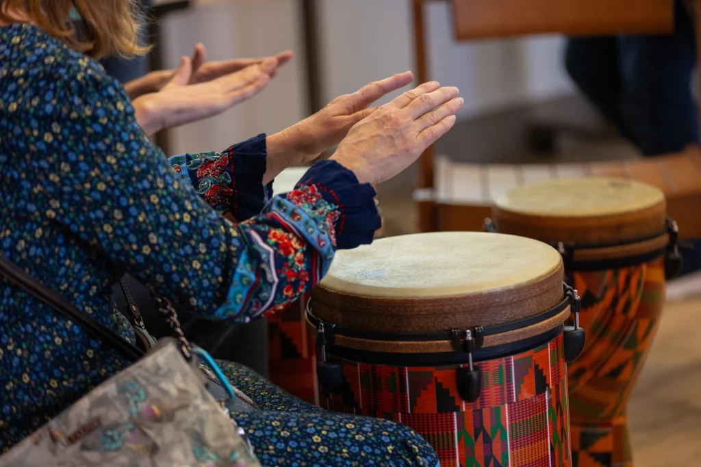 A person in a patterned dress, perhaps part of the Carnegie Hall Lullaby Project, plays hand drums with another nearby—both hands mid-air—capturing a joyful moment that new parents from Valparaiso University might cherish.