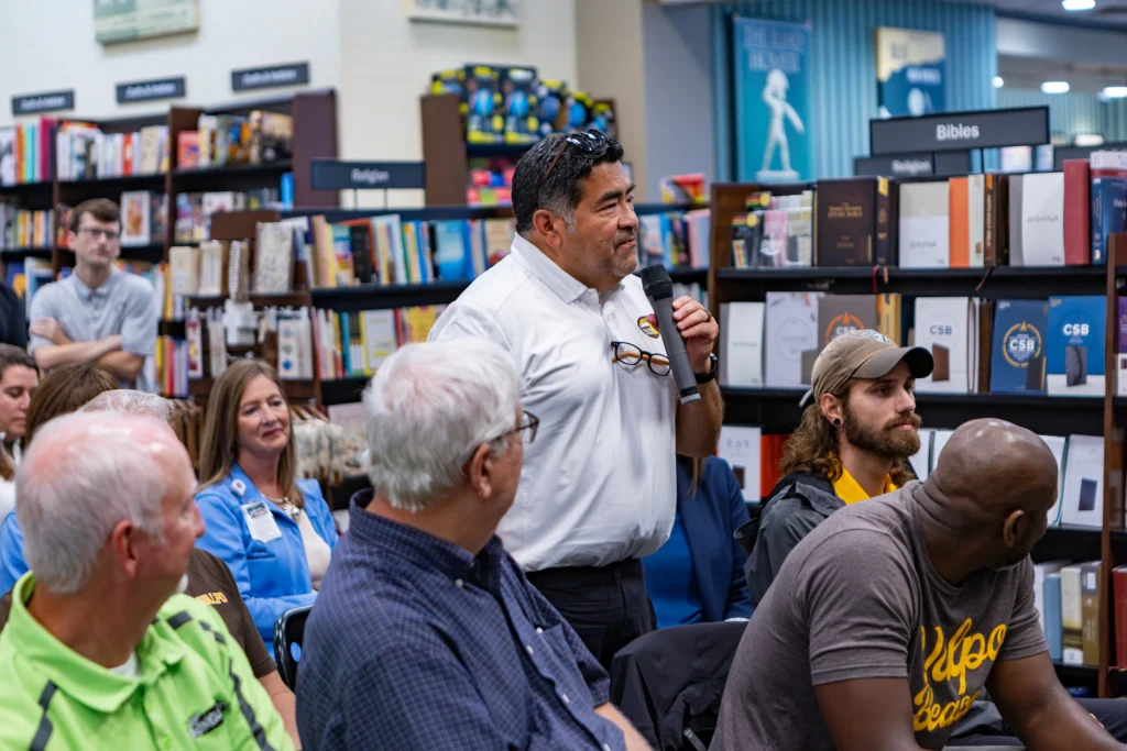 Coach Bob Modesto shares insights from his MBA Journey as he speaks into a microphone amid a seated audience in Valpo’s bookstore religious books section.
