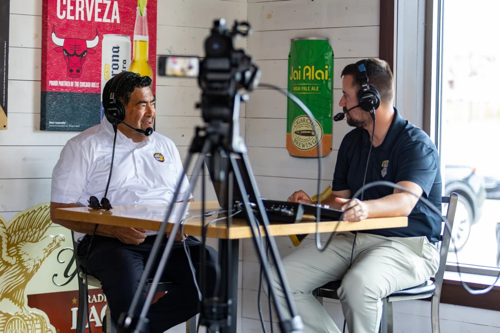 Two men with headsets record a podcast at a table, chatting about their MBA Journey at Valpo, with Coach Bob Modesto making a special appearance; beer signs and a camera sit in the background.