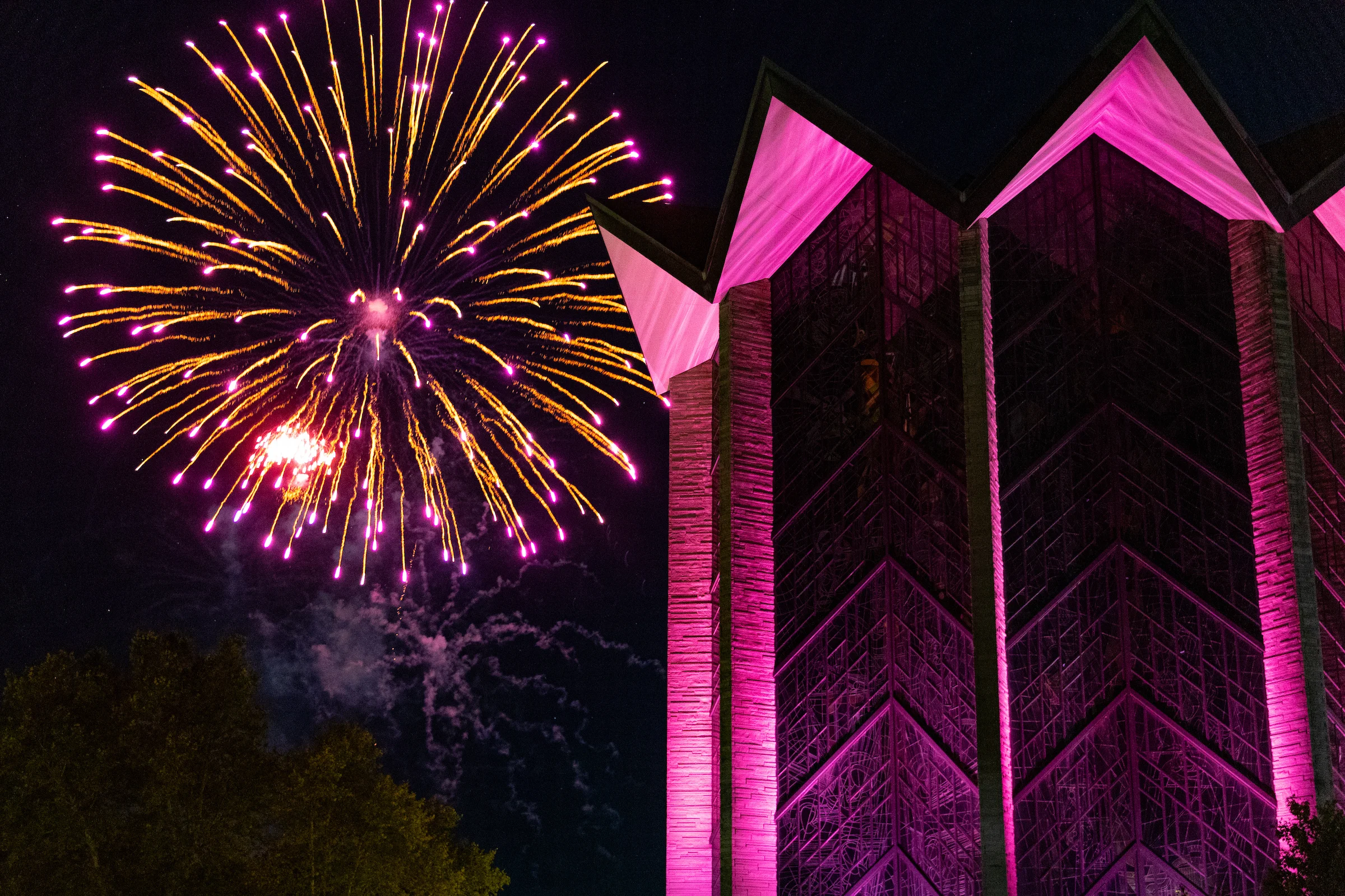 Bright fireworks burst in the night sky next to a modern building lit with vibrant pink lights.