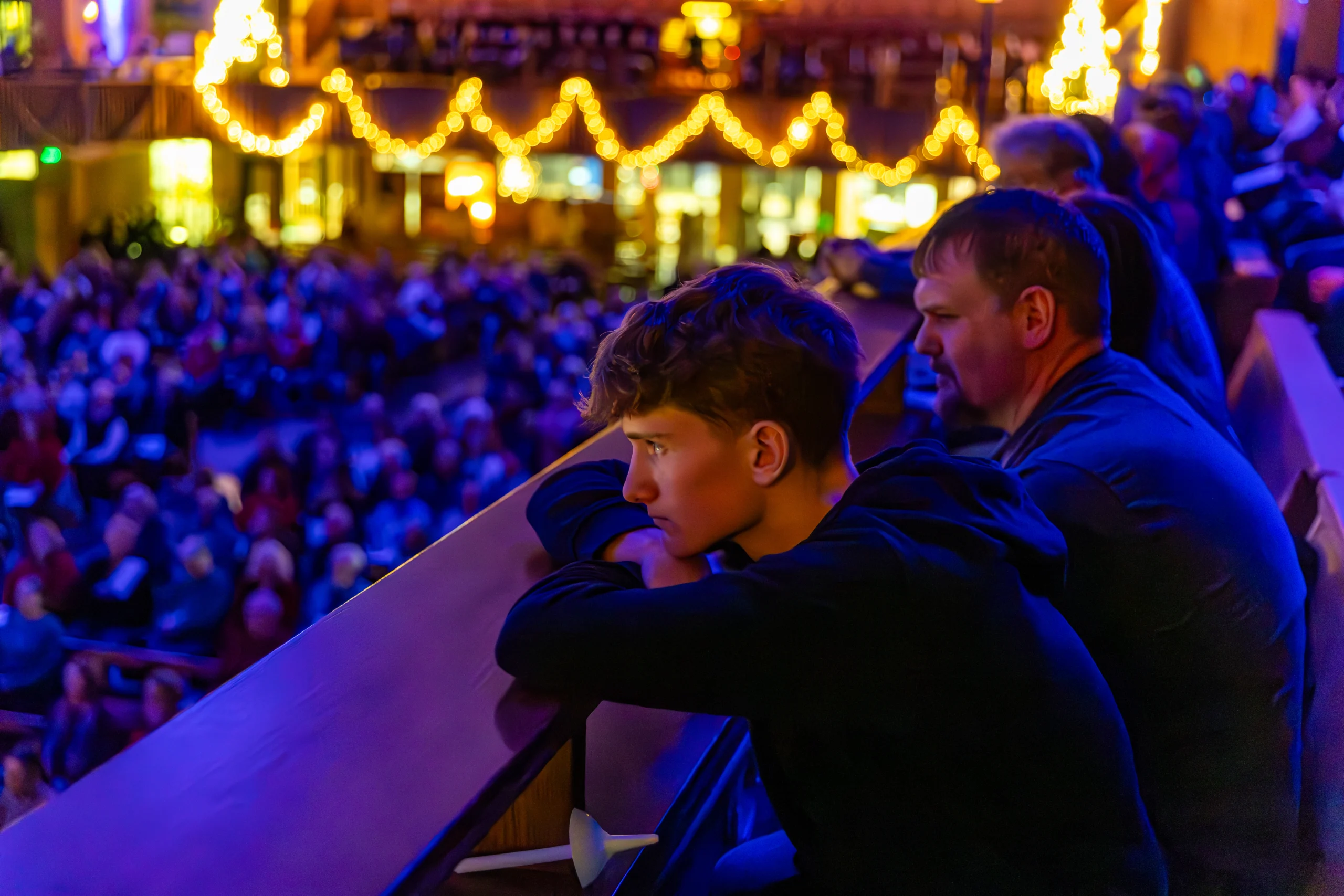 Teen boy leans on balcony railing, immersed in student life as he watches a brightly lit performance with a large audience below.