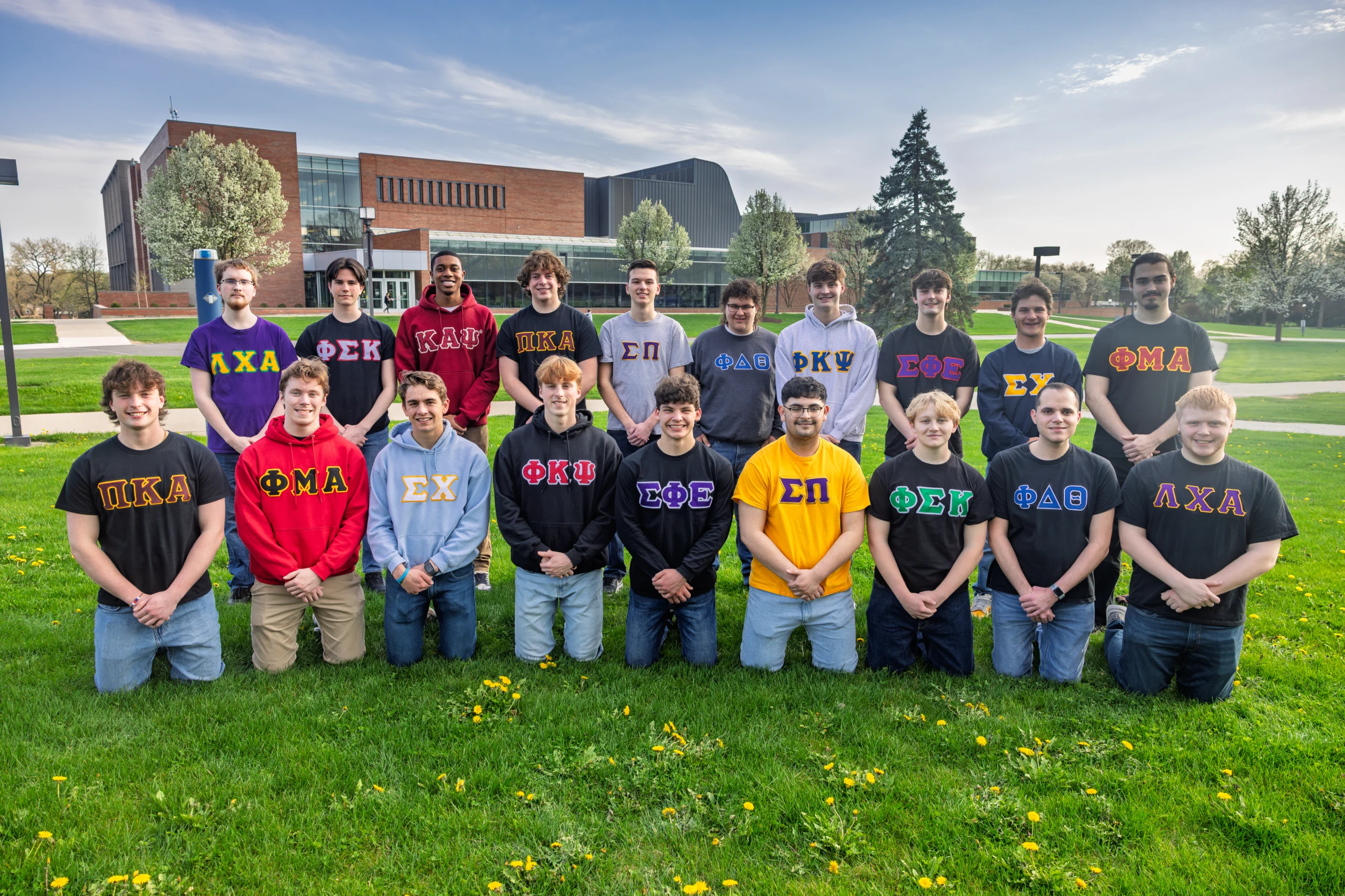 A group of young men wearing Greek letter shirts pose together on a grassy lawn with buildings behind them, capturing a vibrant moment of Student Life.