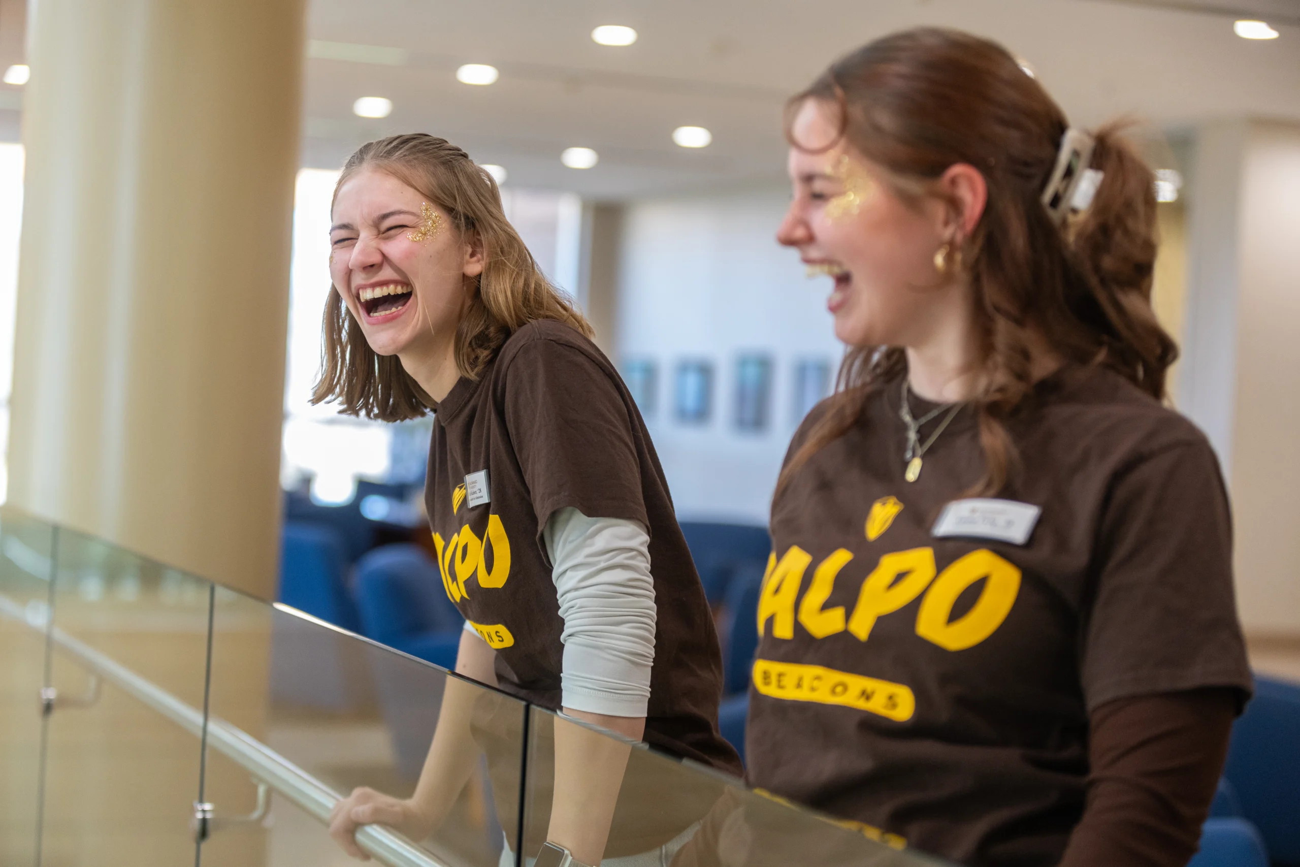 Two people in matching brown ALPO shirts laugh together indoors, leaning on a glass railing, embodying the joyful spirit of student life.