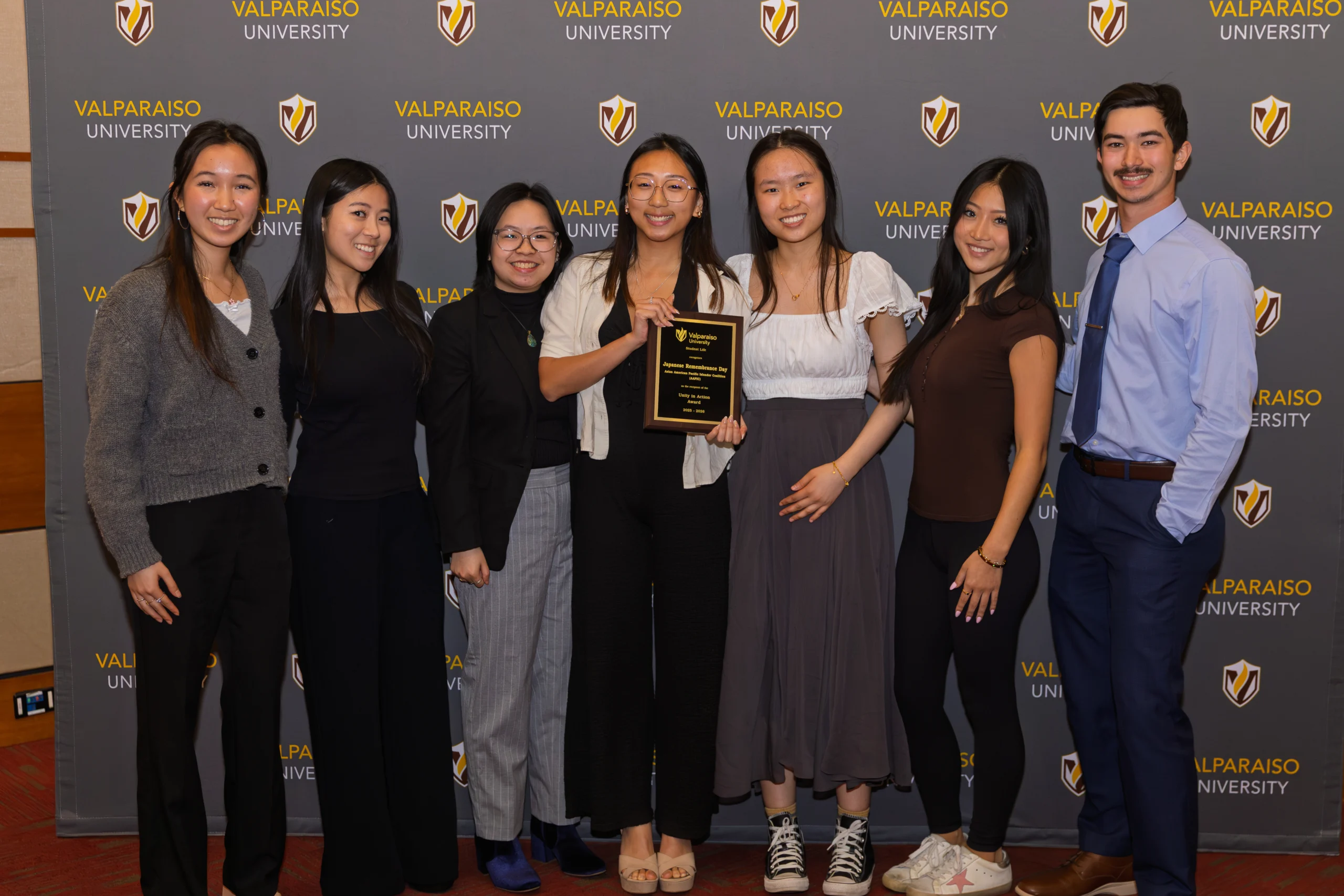 Seven people stand together smiling, with one holding a plaque, in front of a Valparaiso University backdrop—a vibrant moment that highlights the spirit of Campus Life and the achievements made possible through Student Organizations.