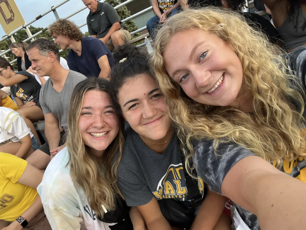 Three smiling young women, including Lauren Bartlett, take a selfie at an outdoor sports event, with a crowd seated behind them beneath the bright Beacon signage.