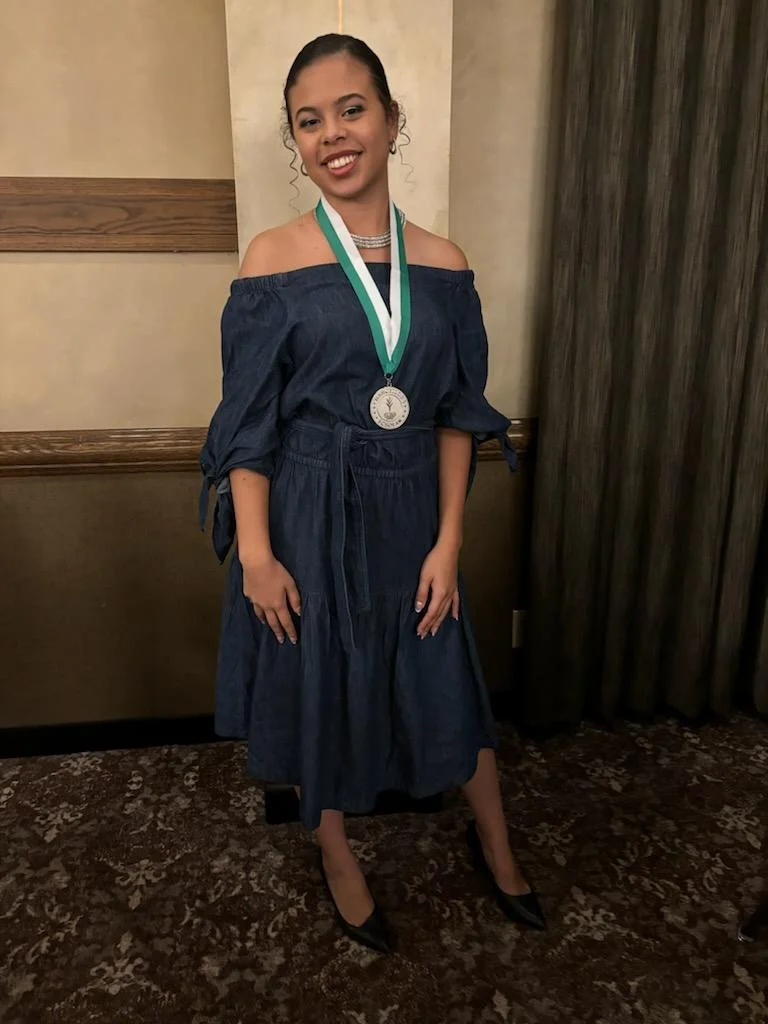 Smiling woman Aria Davis in a denim dress, wearing a medal, stands indoors on a patterned carpet—capturing the spirit of Beacon's Reflection.