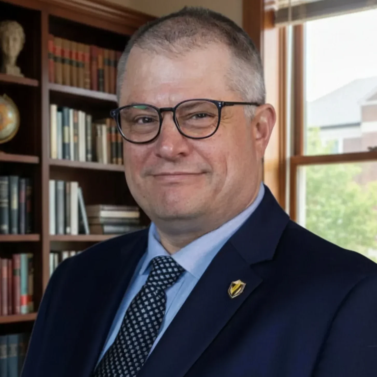 A man in a navy suit and glasses stands in an office with bookshelves and a window in the background.