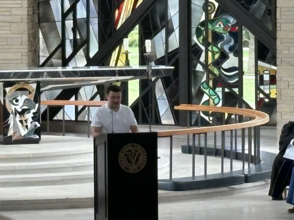 A Valpo student speaks at a podium inside a chapel with stained glass windows and modern art designs, reflecting on their Nuclear Regulatory Commission internship and sharing insights about building a bright future.