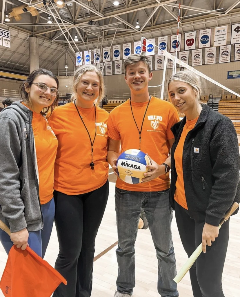 Four smiling people in orange shirts stand in a gym, one holding a Mikasa volleyball, proudly representing Beacon with Lauren Bartlett, M.S., among them.