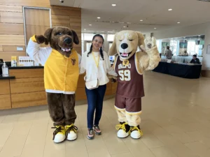 Aria Davis smiles between two dog mascots in yellow and brown outfits inside a modern building lobby, capturing a cheerful moment for Beacon's Reflection.