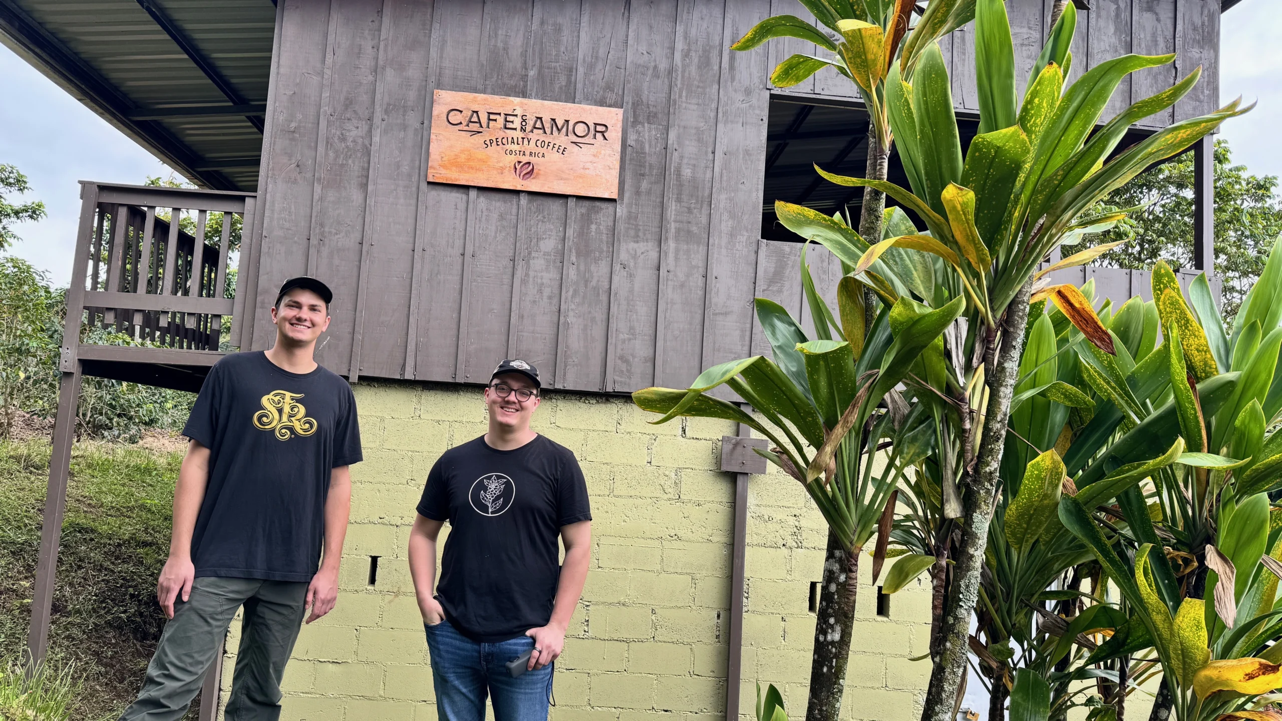Two people stand smiling in front of a Café Amor sign on a wooden coffee shop, with plants to the right—serving as a welcoming beacon for coffee lovers.