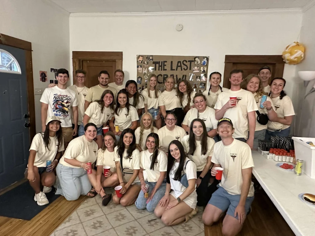 A group of young adults in matching shirts pose and smile together at a party in a living room, celebrating their recent achievements at M.S. Beacon with Lauren Bartlett.