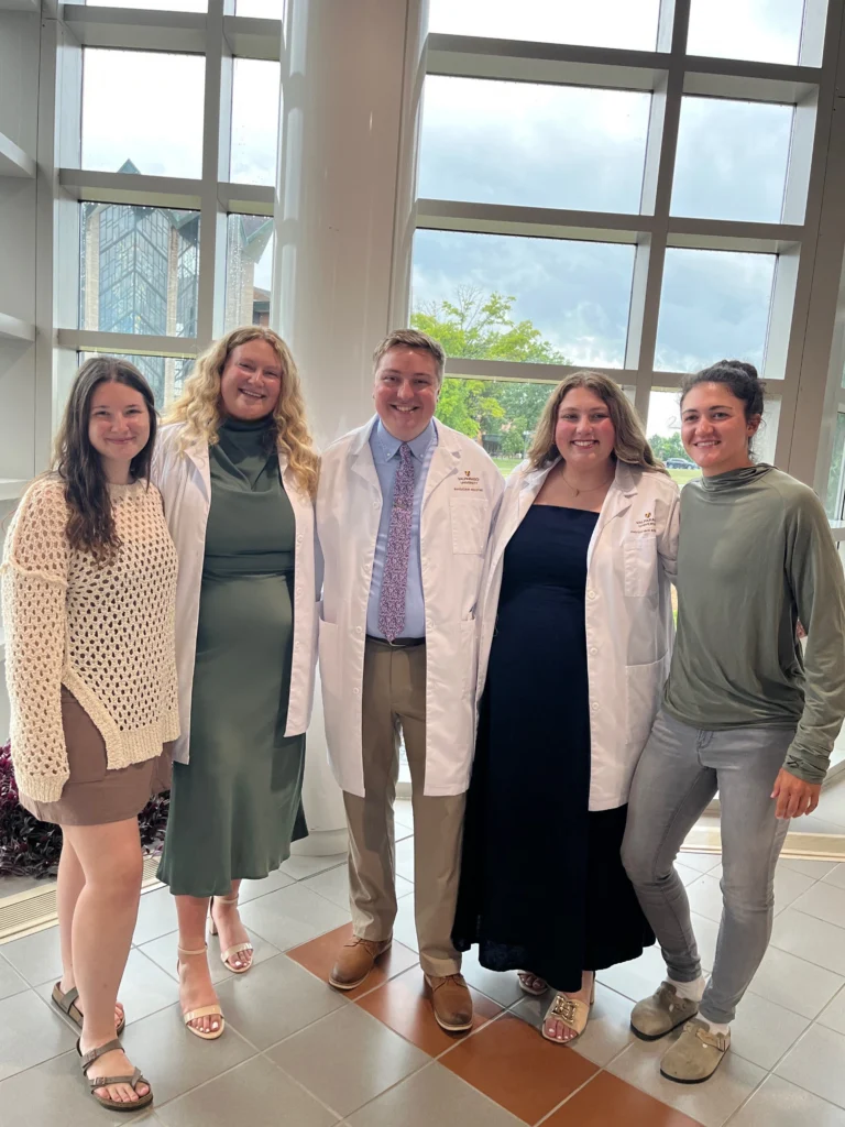 Five people stand indoors, two in white coats, all smiling with large windows in the background—Lauren Bartlett stands among them, embodying the spirit of a Beacon for excellence and teamwork.