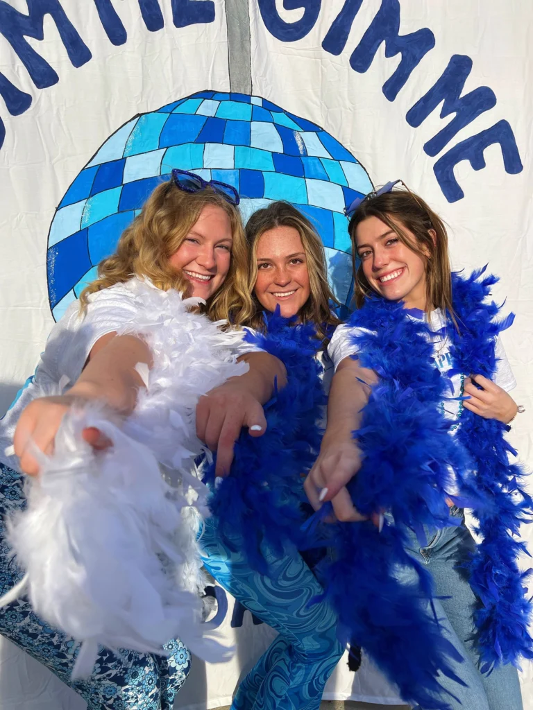 Three smiling women in feather boas pose in front of a blue disco ball backdrop in bright sunlight, radiating joy like a beacon.