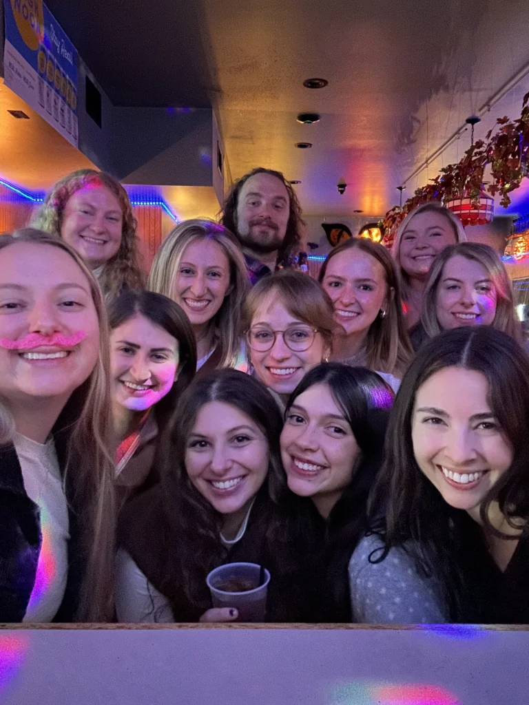 A group of people, including Lauren Bartlett, M.S., are smiling together in a colorful, festive indoor setting with bright lights, creating a true beacon of joy and celebration.