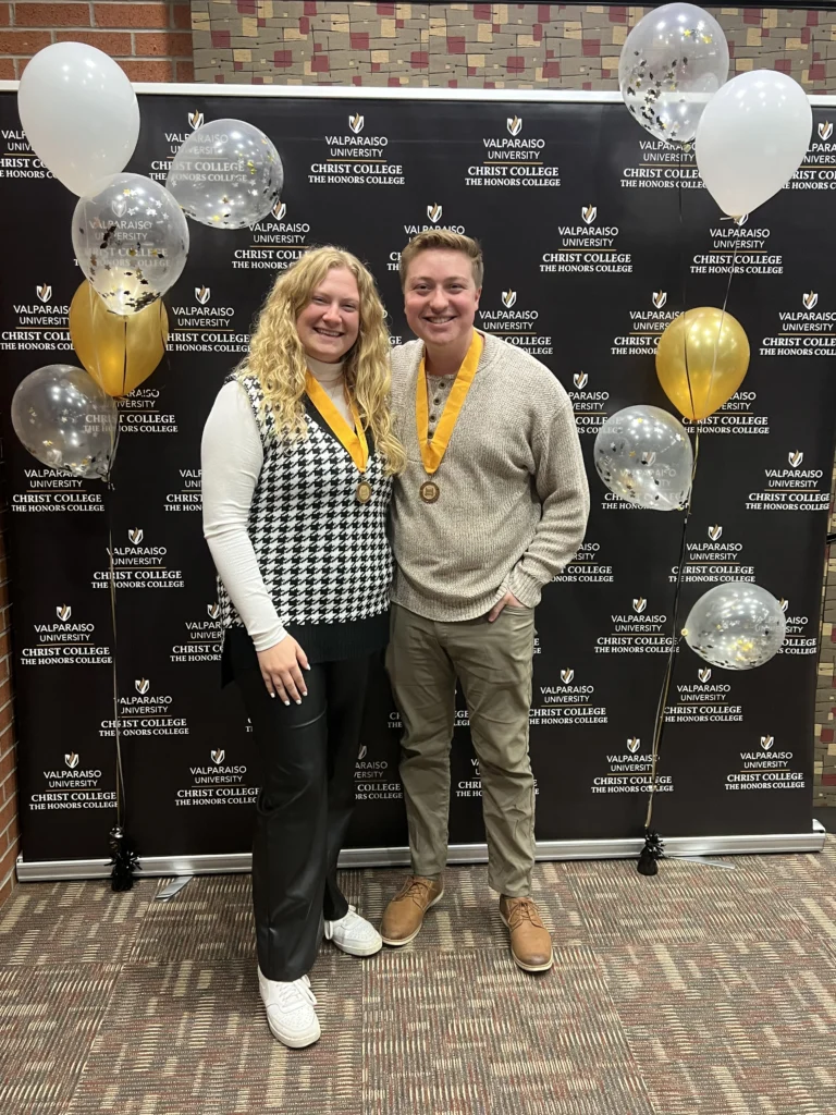 Two people with medals pose, smiling, in front of a Christ College event backdrop with balloons, celebrating their achievements as part of the Beacon program.
