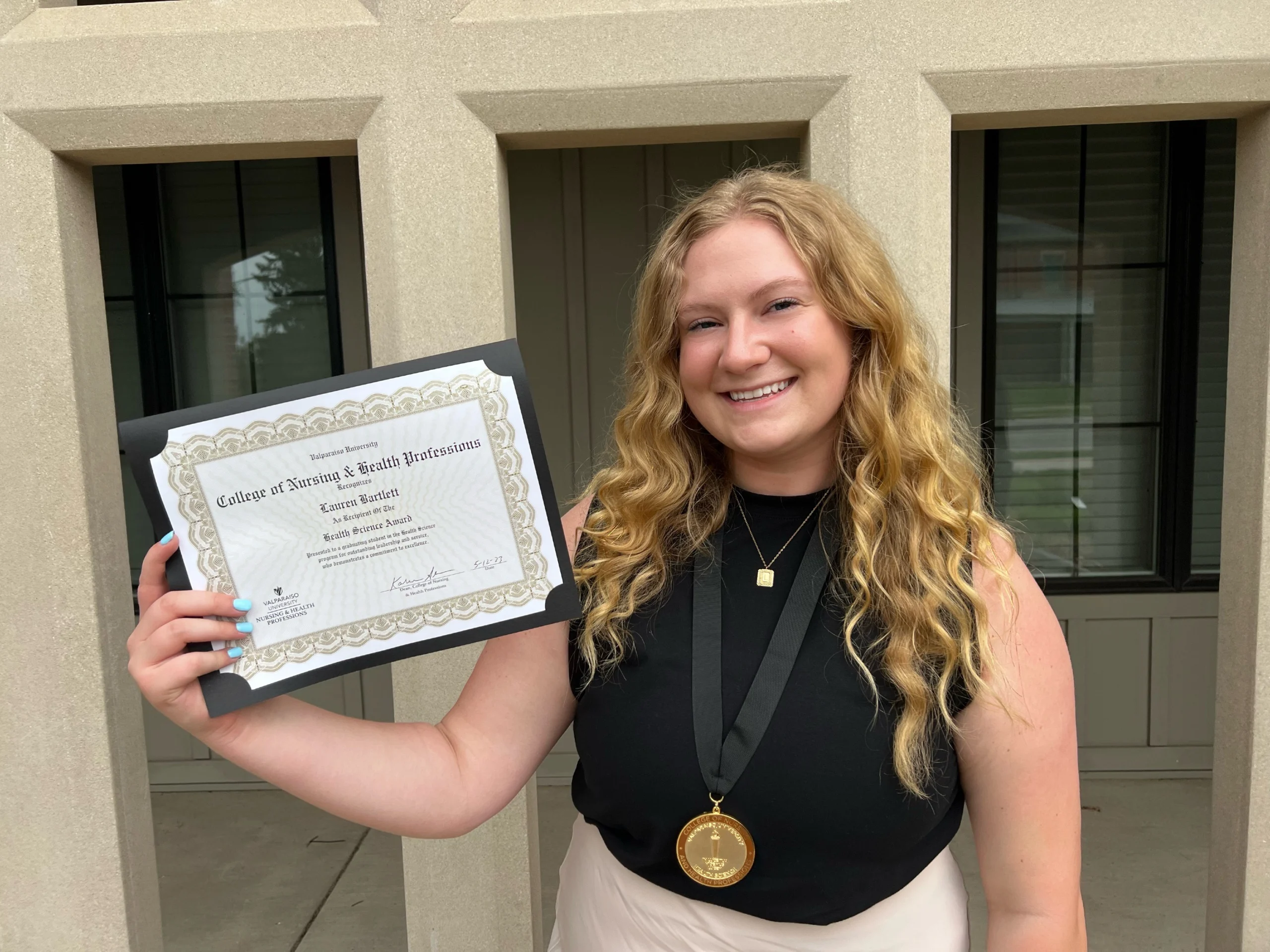 Smiling woman, Lauren Bartlett, holding a certificate and wearing a medal, stands outside a building with beige columns, proudly celebrating her M.S. achievement.