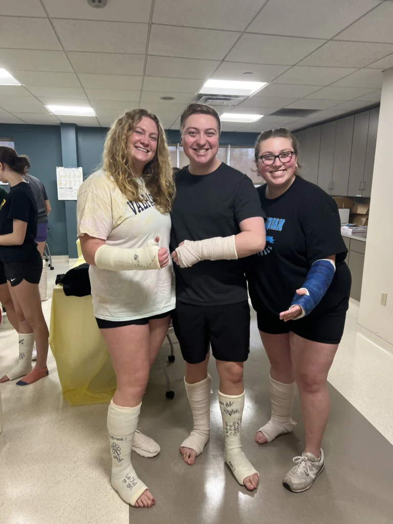 Three smiling people with arm and leg casts pose together in a brightly lit room with medical supplies at Beacon, MS, alongside Lauren Bartlett.