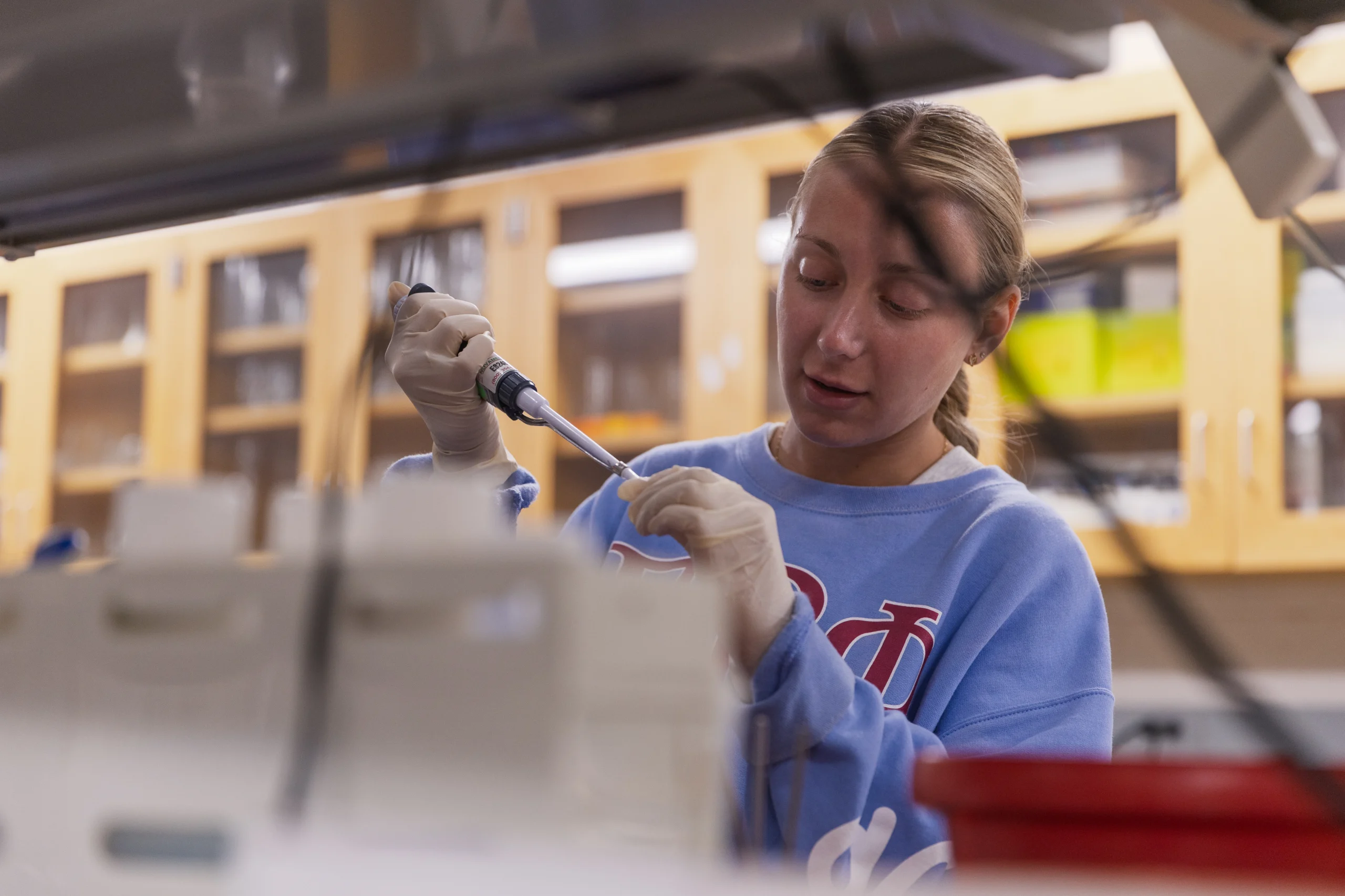 A person in a lab uses a pipette while wearing gloves and a blue sweatshirt, surrounded by lab equipment, as they carefully examine samples of fungus for a research project connected to Valparaiso University.