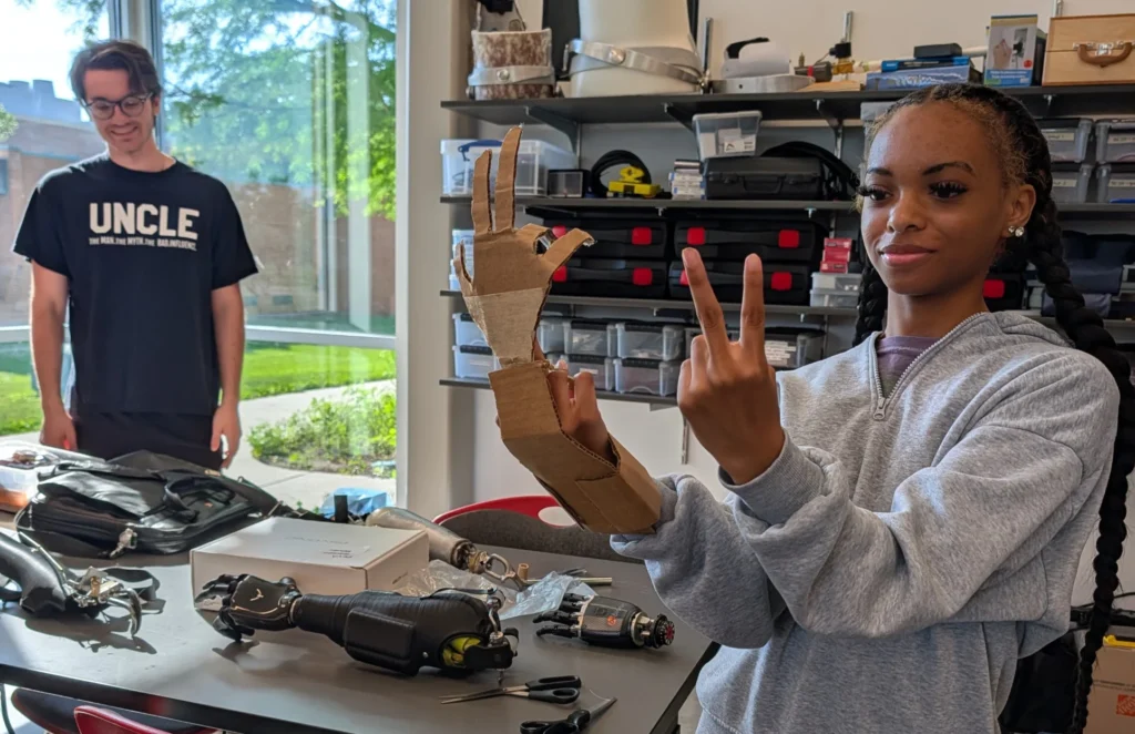 At the Beacon Academy Engineering Camp at Valparaiso University, high school students showcase their creativity as one teen proudly displays a cardboard prosthetic hand in the workshop, while another observes from a table covered with tools.