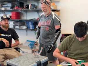 Three men are building robotics at a table; one man, a participant in the Beacon Academy Engineering Camp at Valparaiso University, has prosthetic arms and is smiling at the others.