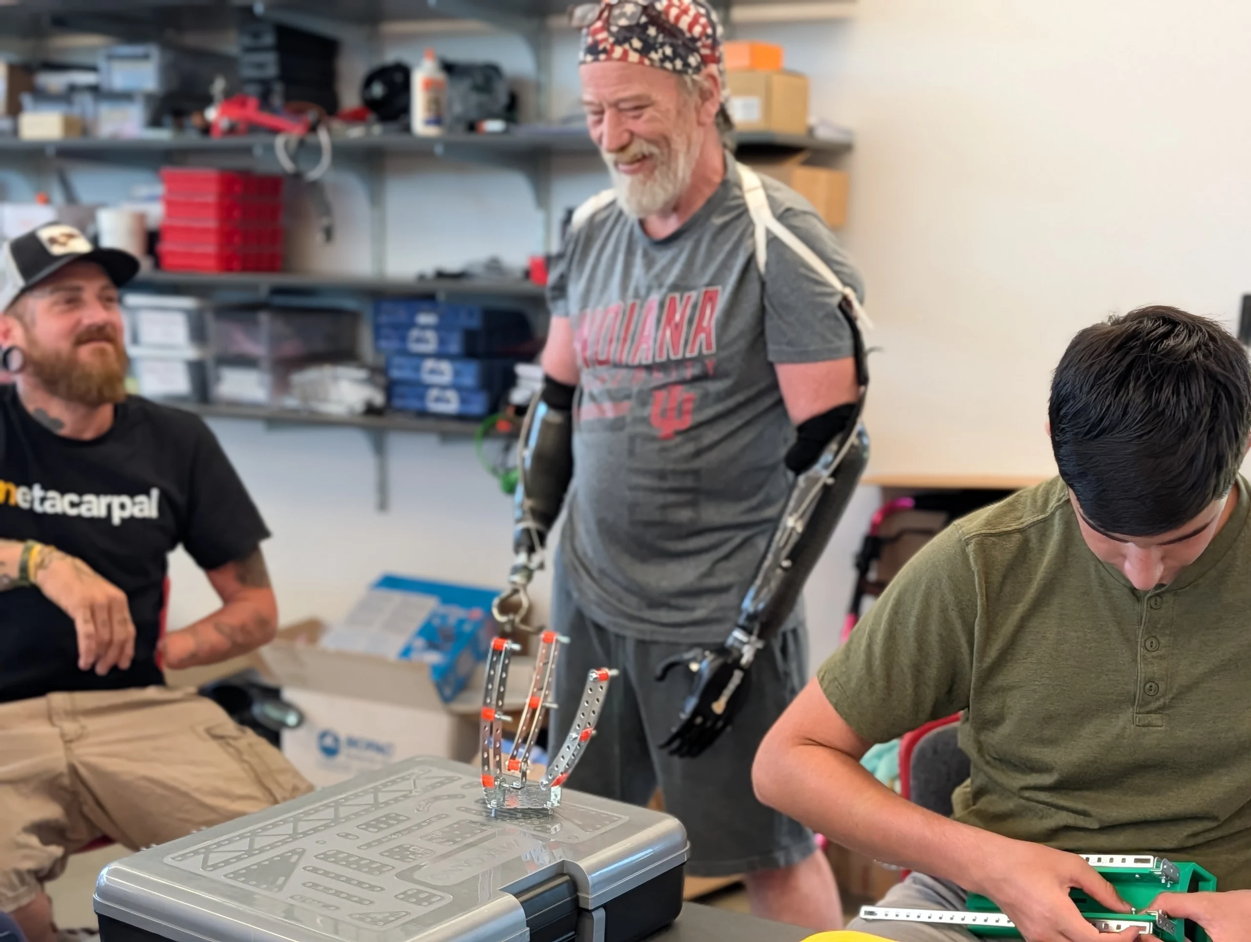 Three men are building robotics at a table; one man, a participant in the Beacon Academy Engineering Camp at Valparaiso University, has prosthetic arms and is smiling at the others.