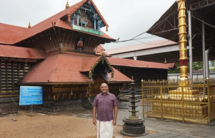 A man in traditional attire stands in front of a South Indian temple with red roofs and golden pillars, embodying the spirit captured by ethnographer George Pati in his Valpo studies.