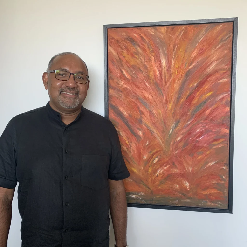 Professor George Pati, chair of the Religious Studies Department at Valparaiso University, stands smiling in a black shirt next to one of his paintings hanging in his office, a vibrant, abstract orange and red piece.