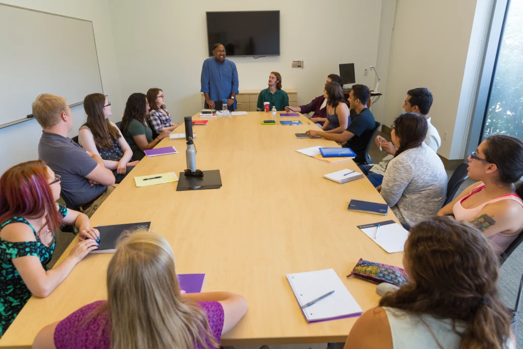 A group of people sit around a conference table, listening as ethnographer George Pati stands at the front of the room, teaching and leading the discussion.